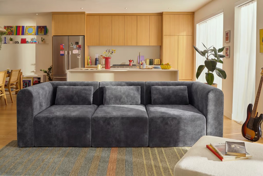 Gray three-seat sofa with separate cushions in a bright shared apartment living room and open kitchen.