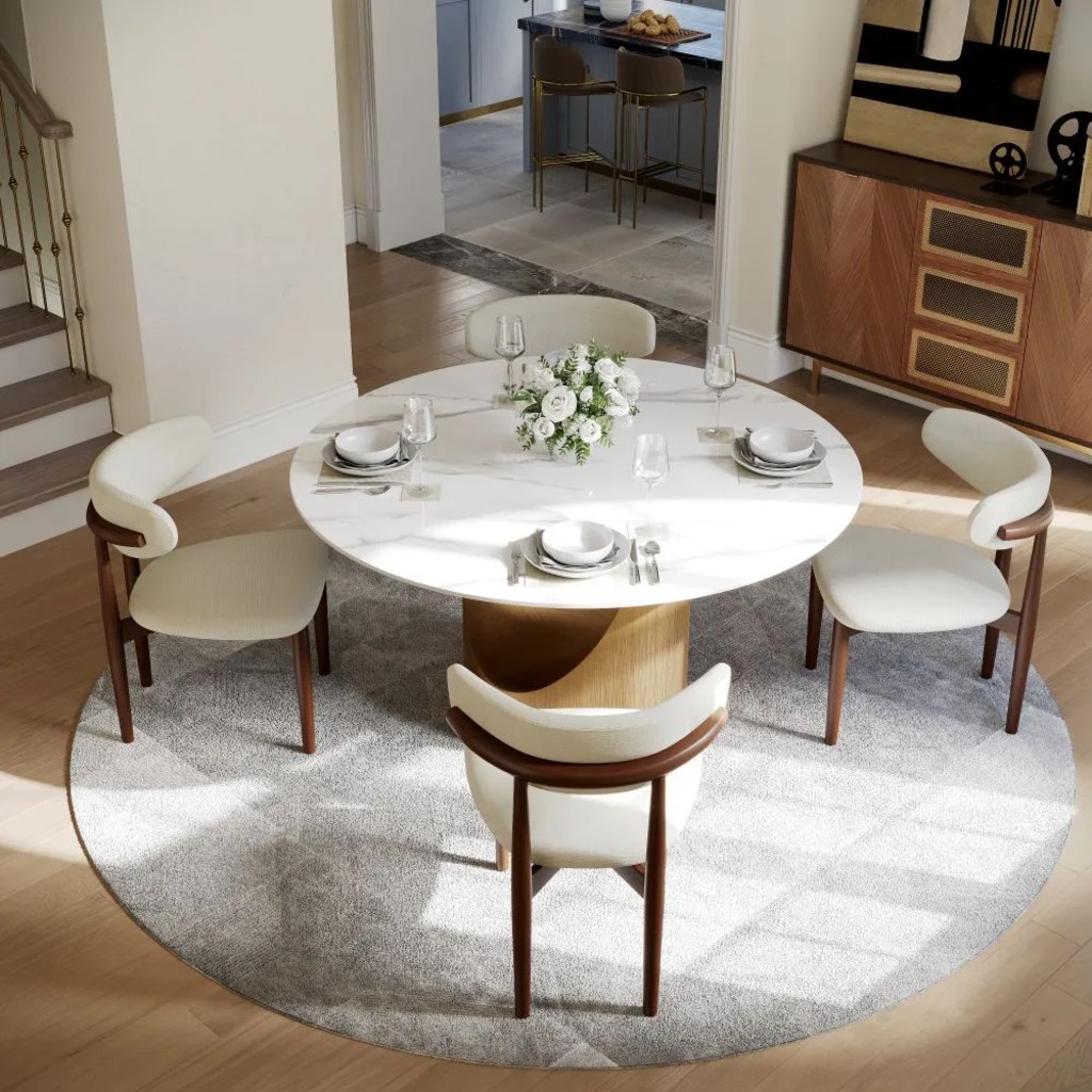 Round white dining table set for four with upholstered chairs on a circular gray rug in a bright modern dining area.