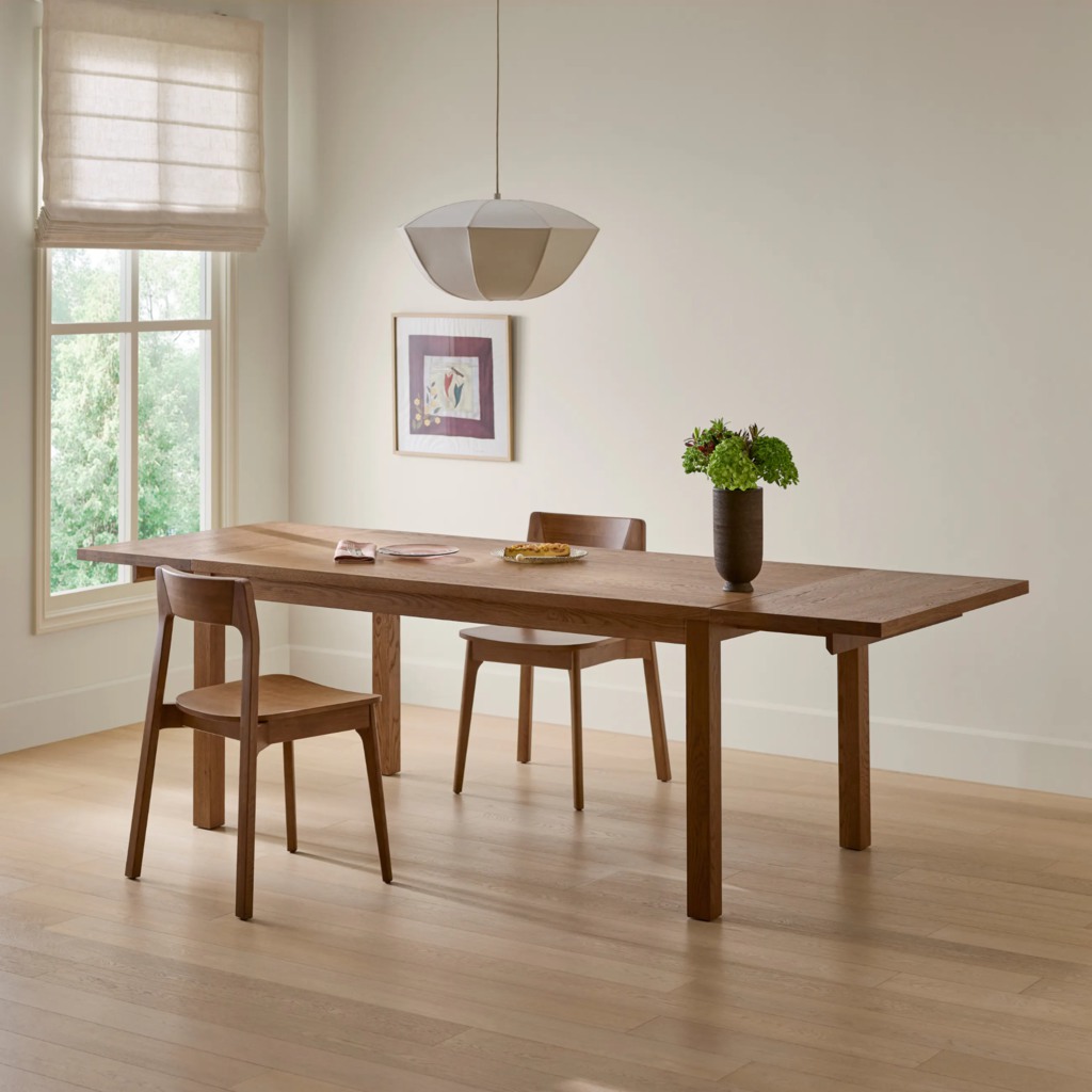 Long rectangular wooden dining table with four corner legs and open space beneath in a bright minimalist dining room.