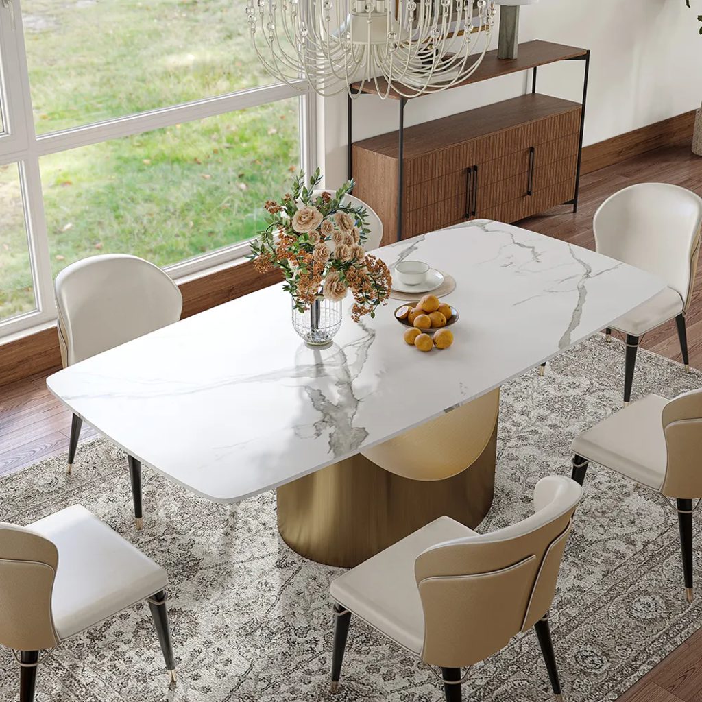 White marble-look dining table with six beige chairs and a gold pedestal base in a bright modern dining room.