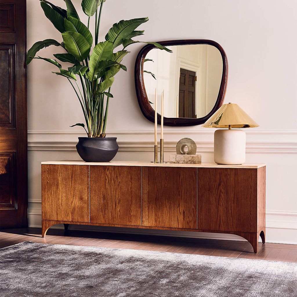 Walnut media console with four closed cabinet doors, styled with a mirror, table lamp, and large indoor plant.