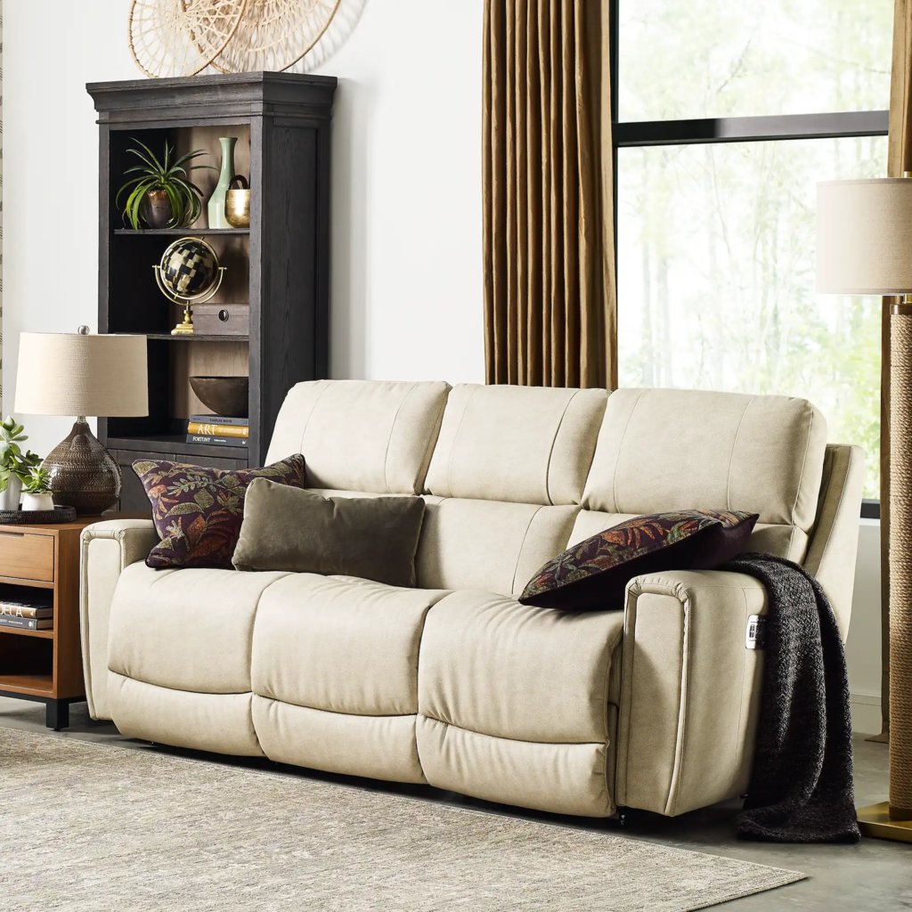 Beige three-seat reclining sofa with pillows and a throw in a bright reading room with bookshelves and floor lamps.