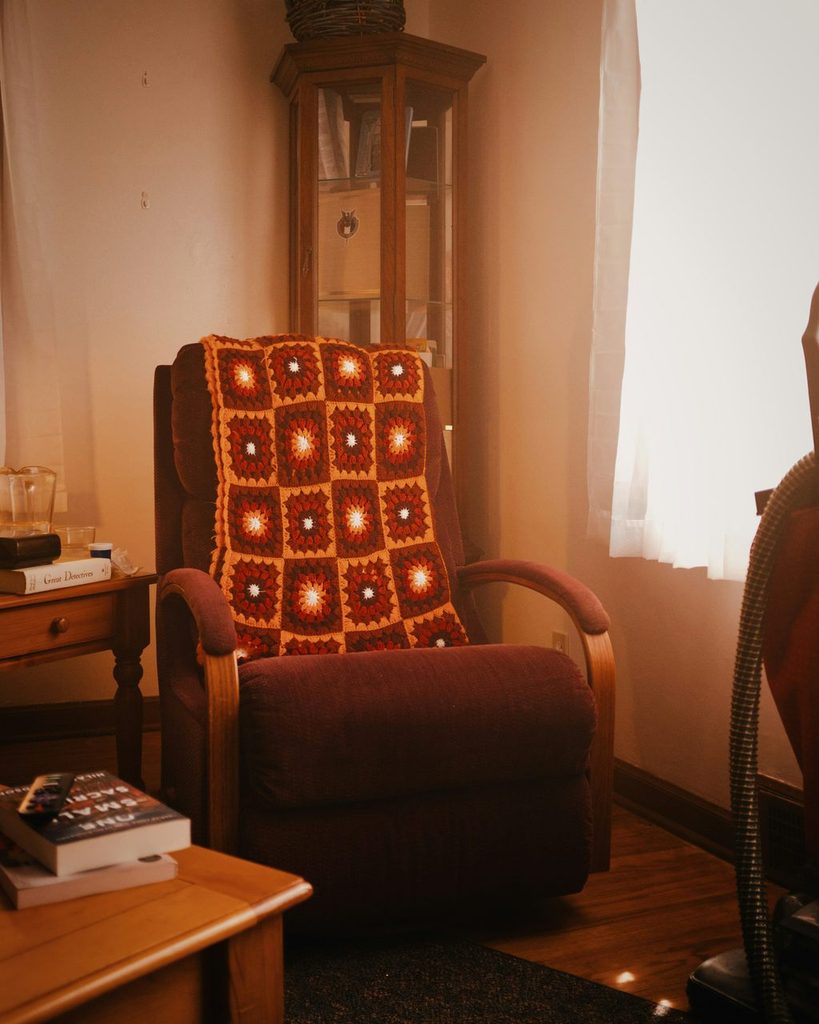 Brown recliner with a crocheted throw beside a window in a warm-toned living room.