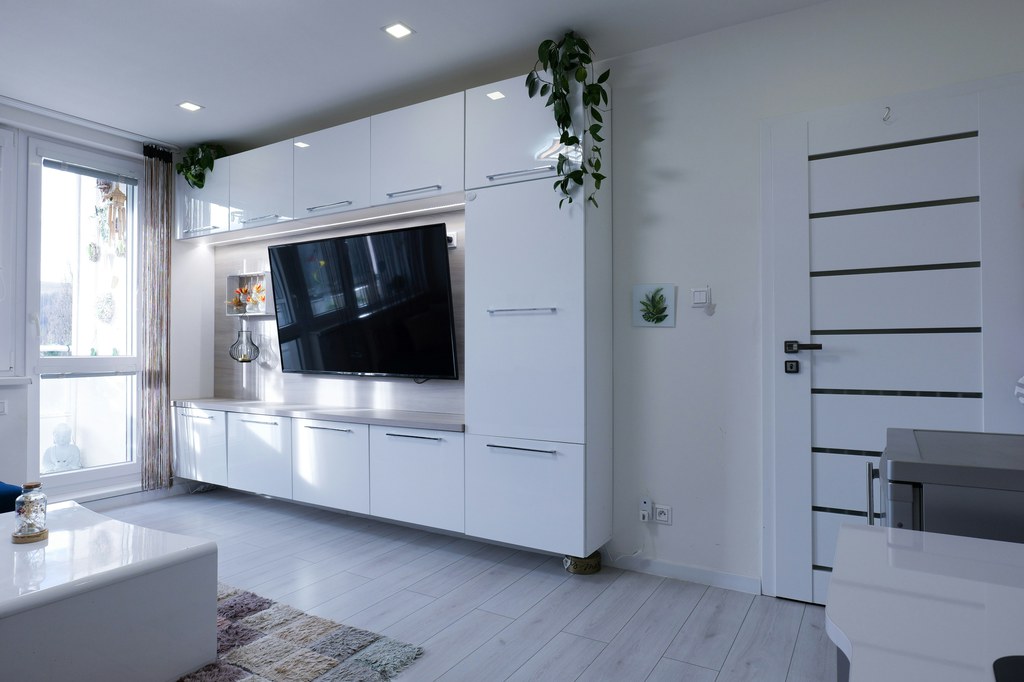 Modern shared apartment living room with a wall-mounted TV, extensive white storage cabinets, and clear floor space.