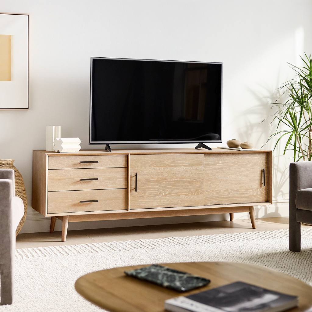 Light wood mid-century media console with three drawers and sliding cabinet doors supporting a flat-screen TV in a bright living room.