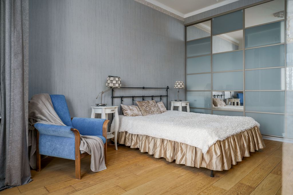 Blue upholstered bedroom reading chair beside a bed with nightstands and mirrored wardrobe.