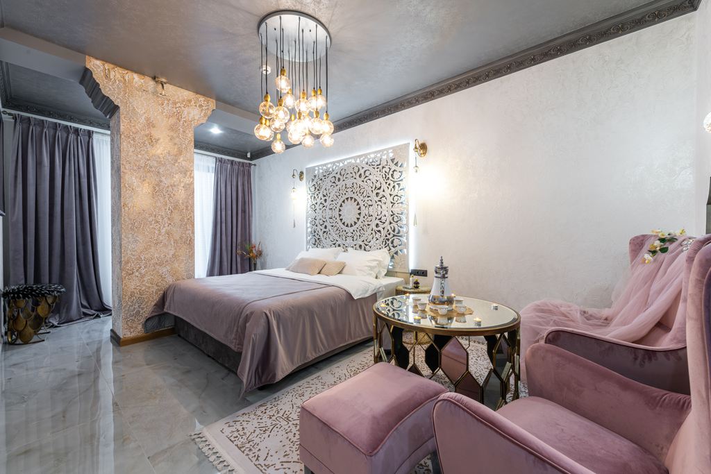 Pink upholstered bedroom armchairs with ottomans beside a bed and round coffee table.