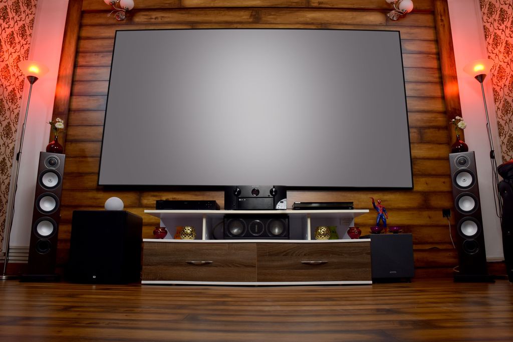 Home theater setup with a large projector screen, floor speakers, center speaker, AV receiver, and media console in a wood-paneled room.