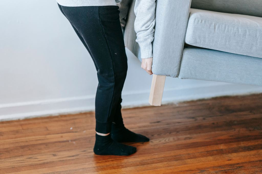 Person lifting a gray sofa to move it safely across hardwood flooring.