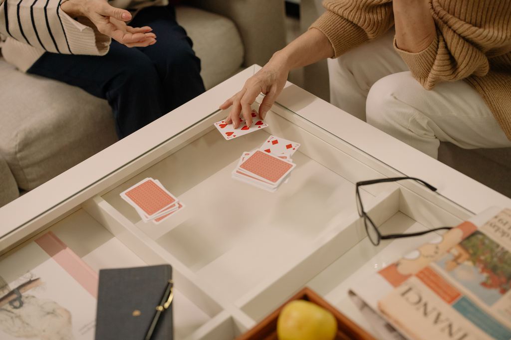Two people playing a card game on a white coffee table in a cozy living room setting with snacks and everyday items nearby.