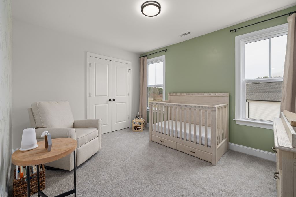 Bright baby nursery with a beige recliner, wooden crib, side table, and soft green accent wall.