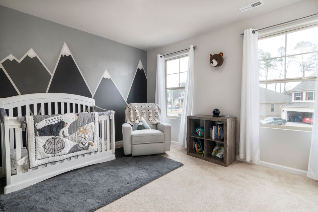 Bright baby nursery with a white crib, gray recliner, bookshelf, and mountain-themed accent wall.