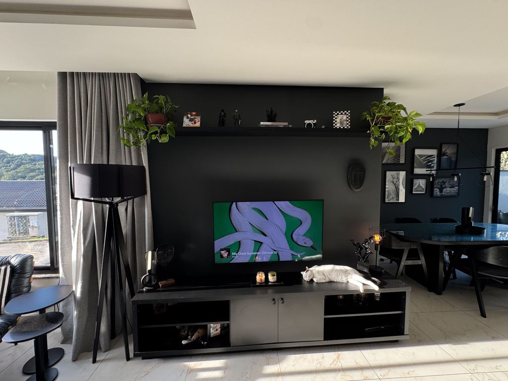 Modern living room with a black media console beneath a wall-mounted TV, surrounded by plants, decor, and dining furniture.