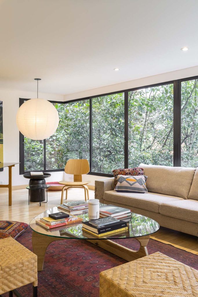 Modern living room with a glass coffee table, beige sofa, lounge chairs, books, rug, and large windows overlooking greenery.