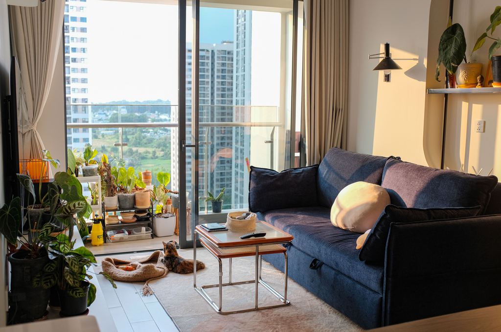 Dark blue sofa in a compact apartment living room with a coffee table, indoor plants, balcony doors, and city views.