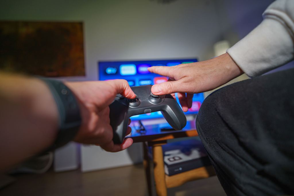 Two people sharing a game controller in front of a TV and media console.