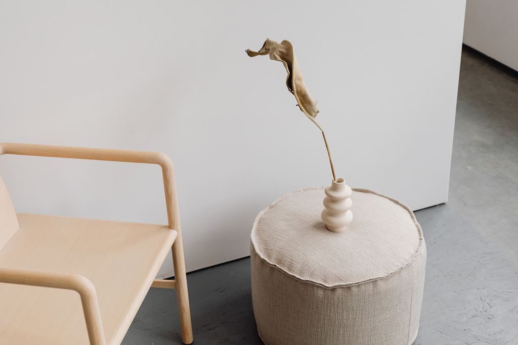 Beige round fabric pouf beside a light wood chair in a minimalist interior.