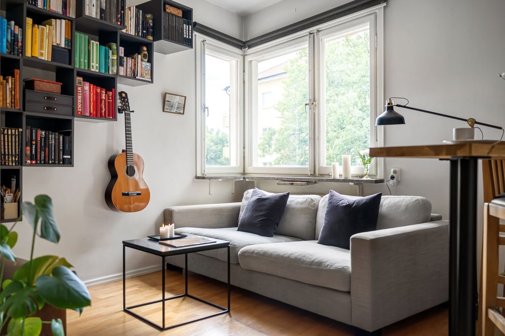 Light gray sofa in a bright reading nook with wall-mounted bookshelves, a guitar, coffee table, and large corner windows.