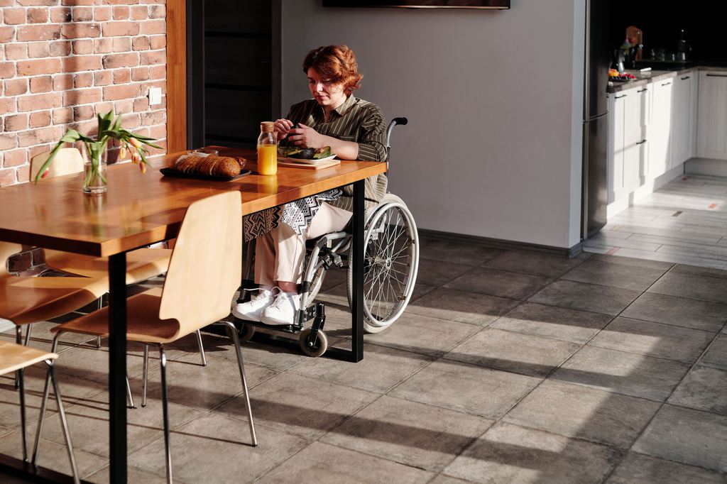 Wheelchair user seated at a wooden dining table with clear floor space in a modern kitchen.