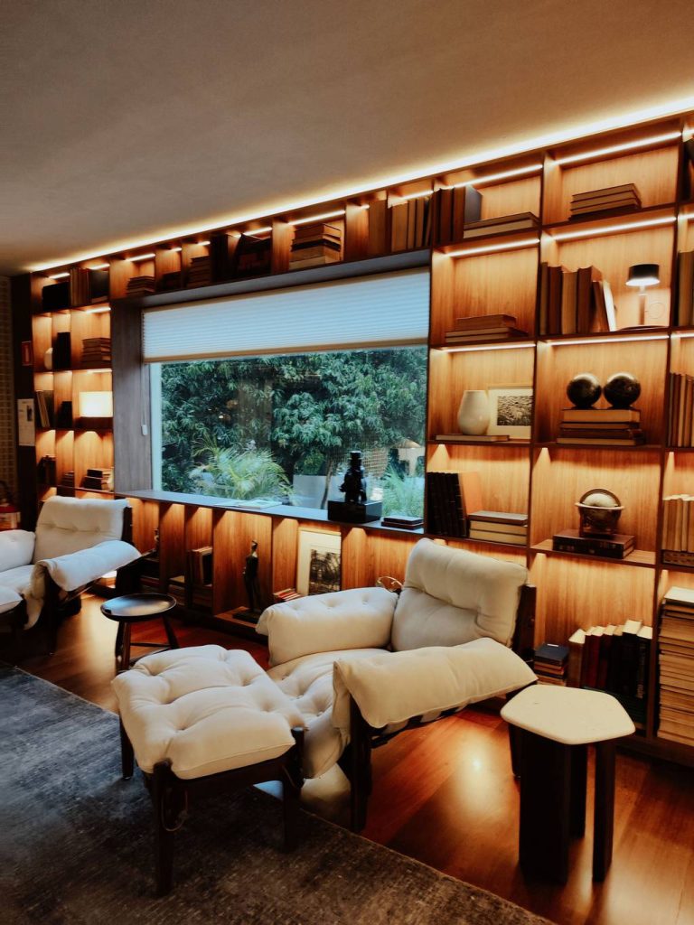 Two cream armchairs with matching ottomans in a warm home library with built-in wooden bookshelves and ambient lighting.