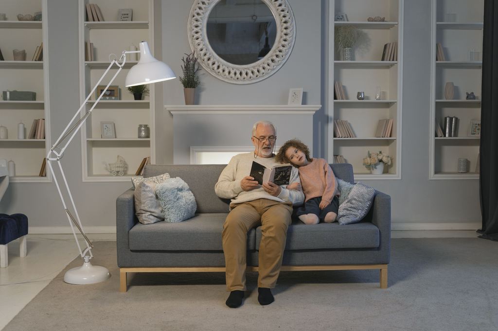 Older man and child reading together on a gray sofa in a cozy living room with bookshelves and a floor lamp.