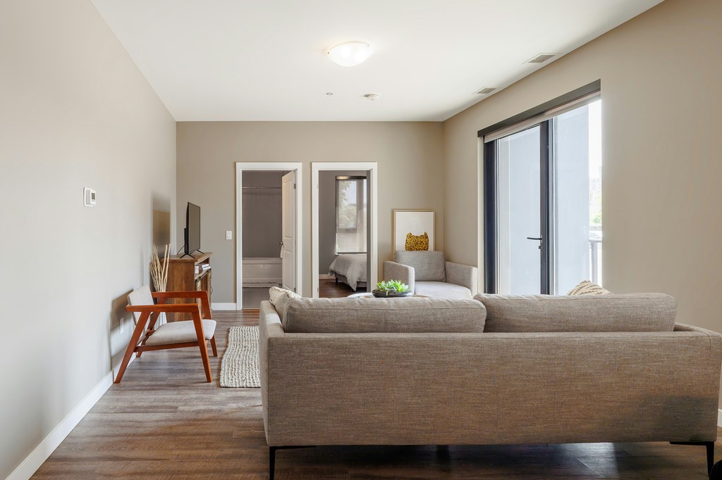 Neutral shared apartment living room with a gray sofa, two armchairs, TV, and clear walkways to adjoining rooms.