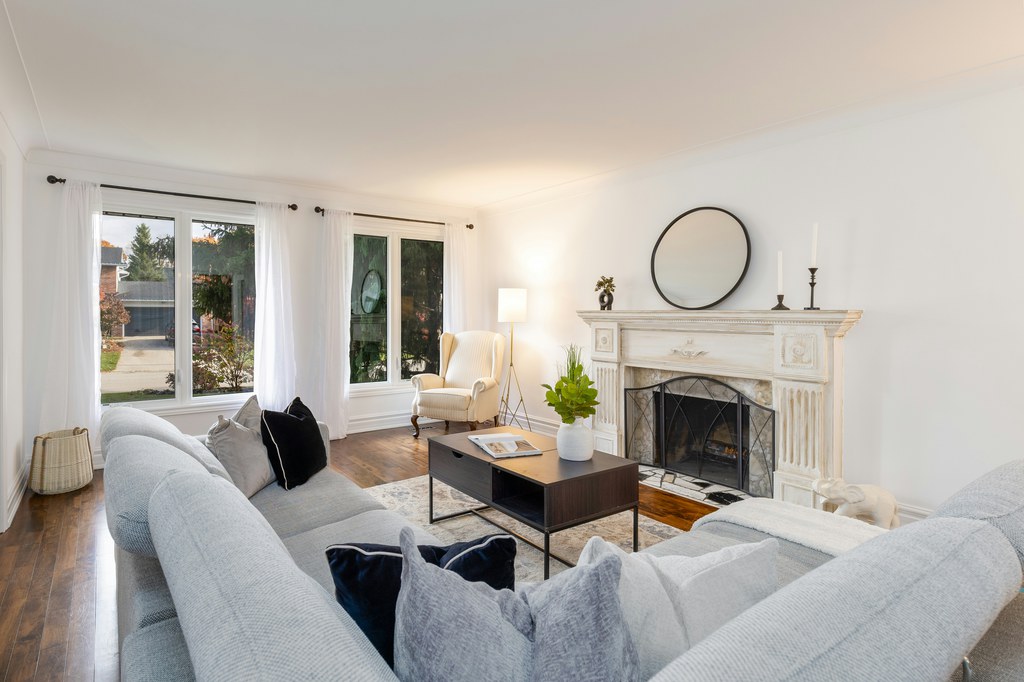 Light and airy living room with gray sofas, sheer white curtains, dark wood flooring, a fireplace, and a round wall mirror.