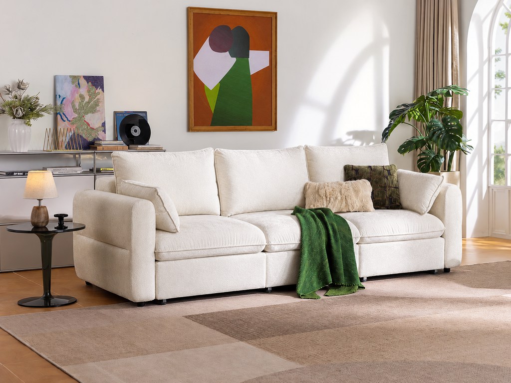 Cream fabric sofa with textured cushions, green throw blanket, and accent pillows in a bright modern living room with abstract wall art, plants, rug, and natural window light.