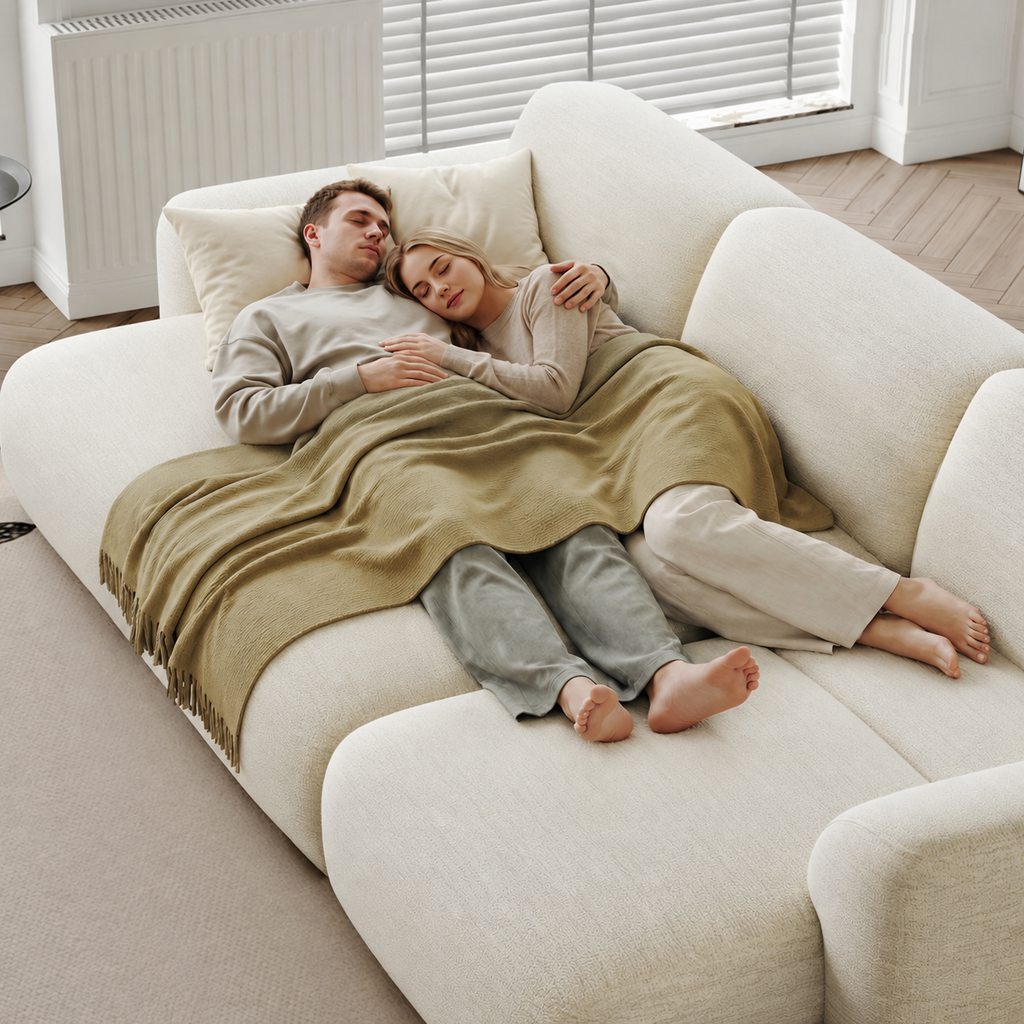 Couple sleeping comfortably on a cream sofa bed under a blanket in a bright living room.