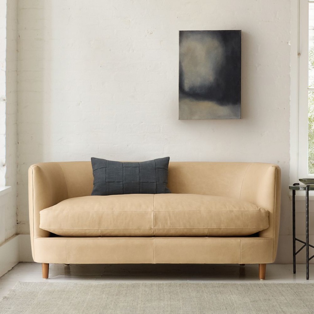 Compact tan high-arm loveseat with a deep seat and single dark throw pillow in a minimalist living room.