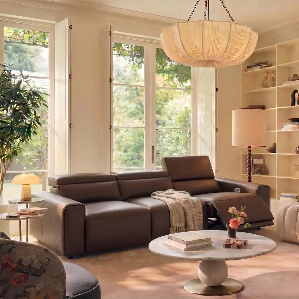 Black reclining sofa with adjustable headrest beside a round coffee table in a sunlit living room.