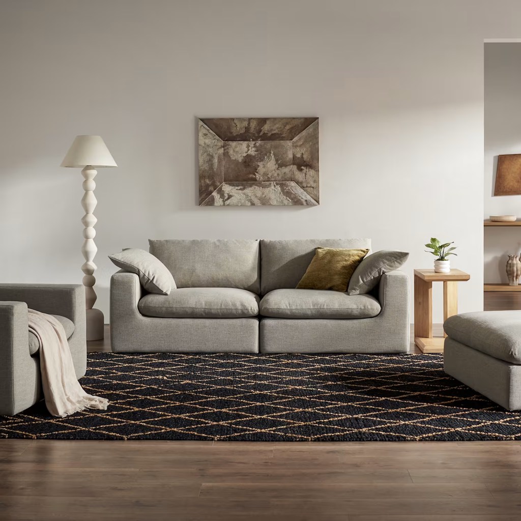 Light gray fabric sofa with loose cushions in a modern living room with neutral decor, wood flooring, and a black patterned rug.