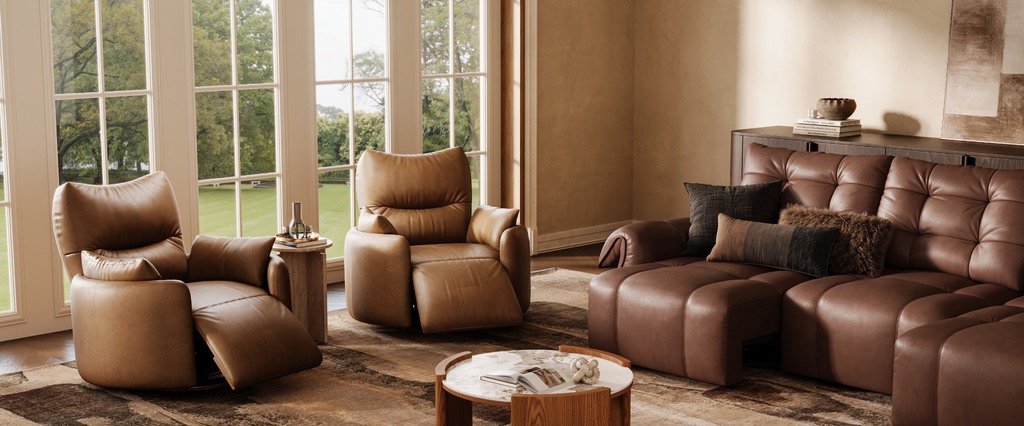 Two tan leather recliners and a brown reclining sofa arranged on a patterned rug in a bright living room.