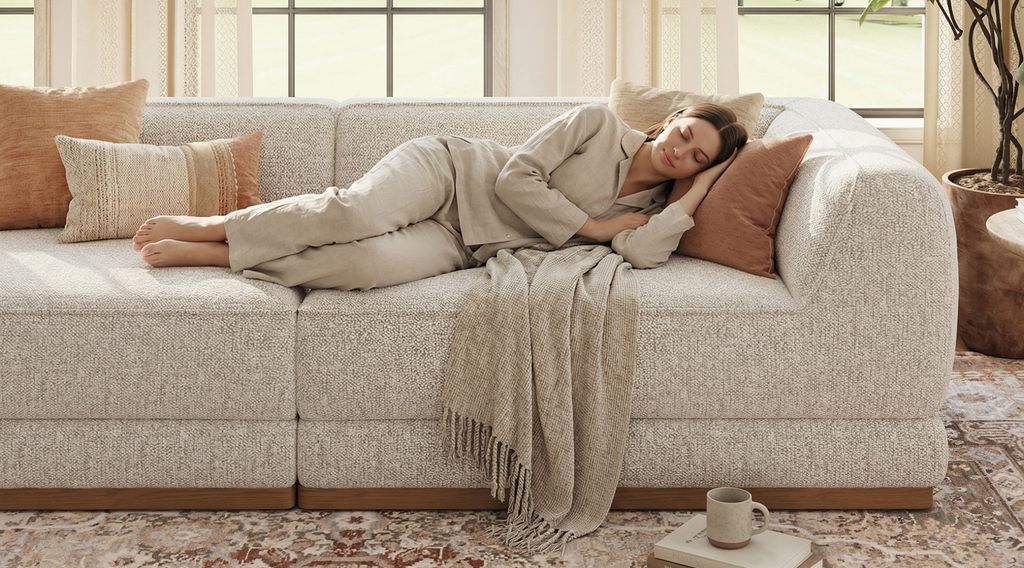 Woman resting across a light beige sofa with throw pillows in a sunlit living room.