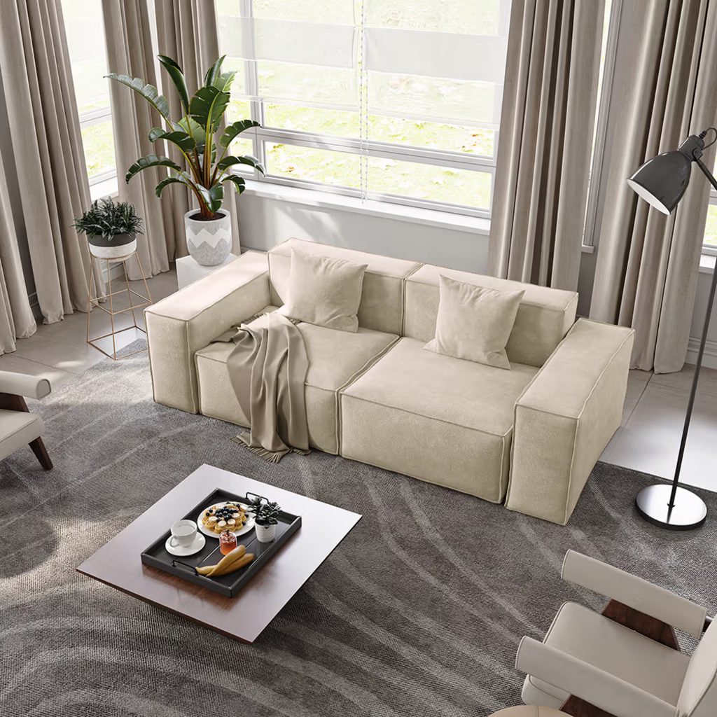 Cream modular sofa with wide armrests and throw pillows in a bright living room with large windows, neutral curtains, and a coffee table.