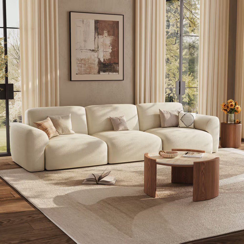 Modern Mediterranean living room with a cream curved sofa, stone-look and walnut coffee table, neutral rug, warm wood flooring, and soft natural light.