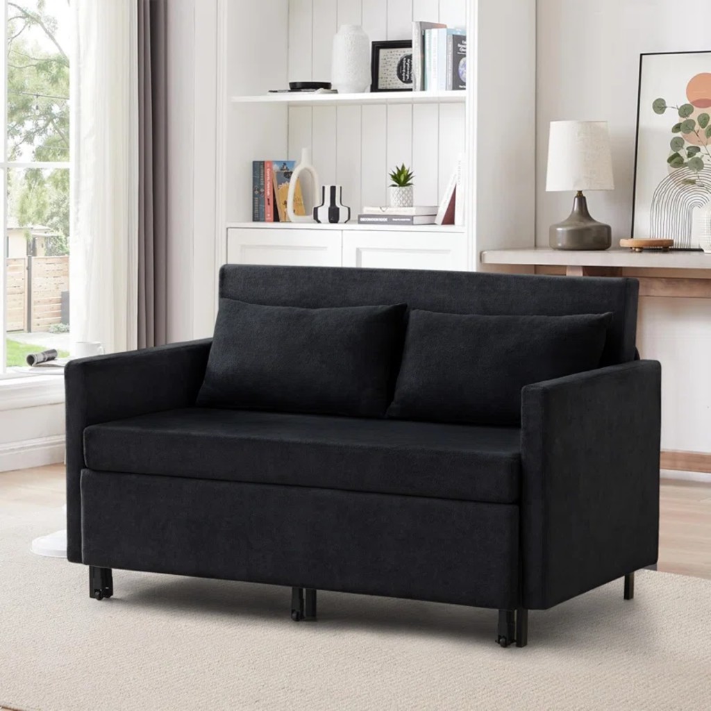Black fabric sleeper sofa with clean lines and storage base design in a modern living room with built-in shelves, lamp, rug, and natural light.