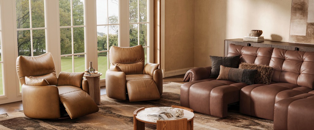 Tan leather recliners and brown leather sofa in a bright living room with warm wood floors and large windows