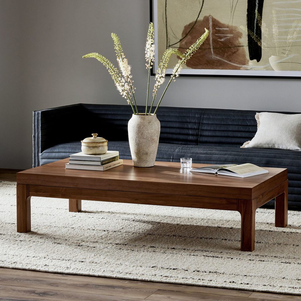 Large rectangular walnut coffee table with books and a vase in front of a black sofa in a modern living room.