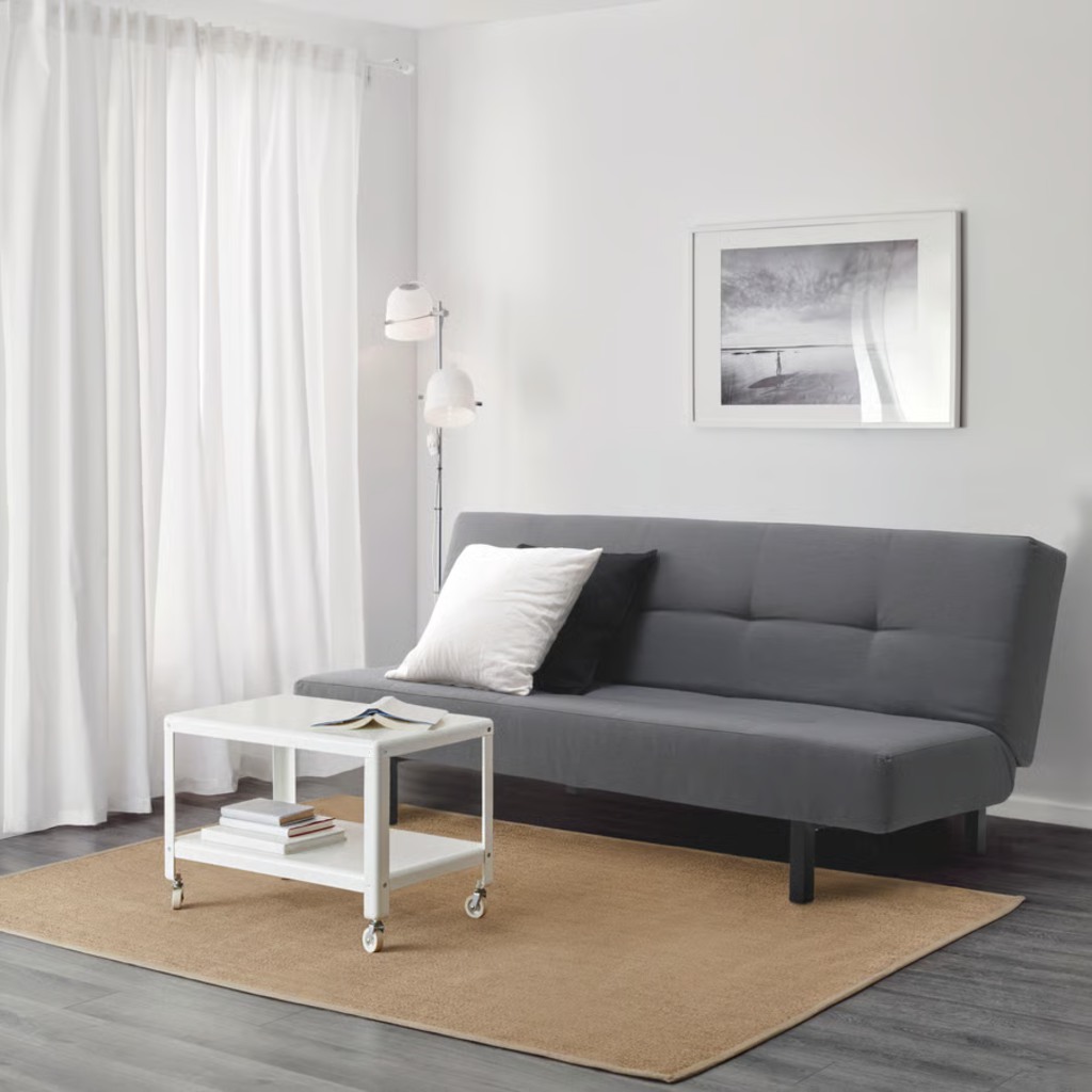 Dark gray sleeper sofa with adjustable backrest and minimalist design in a bright modern living room with white curtains, coffee table, rug, and wall art.