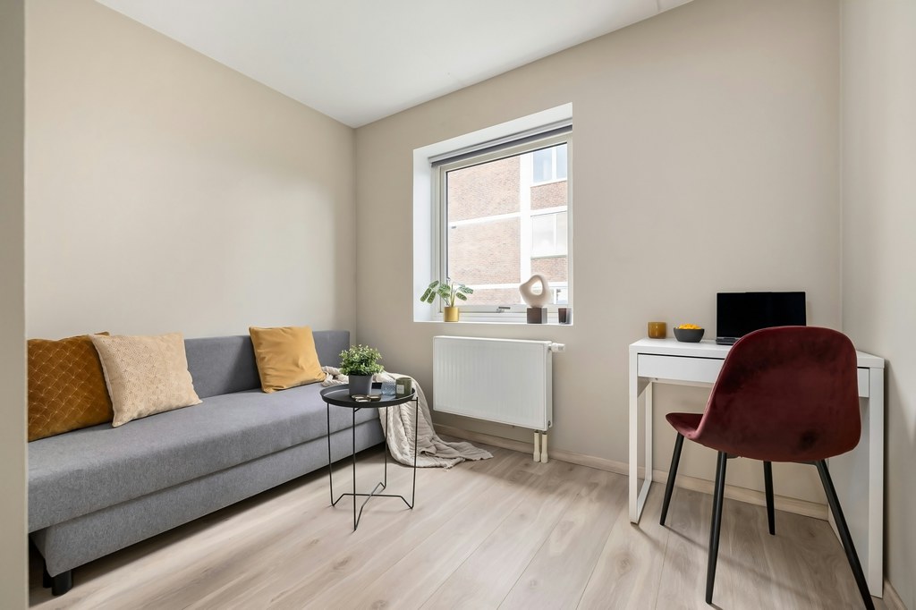 Living room office with a gray sofa, white desk, laptop, and burgundy chair beside a window.