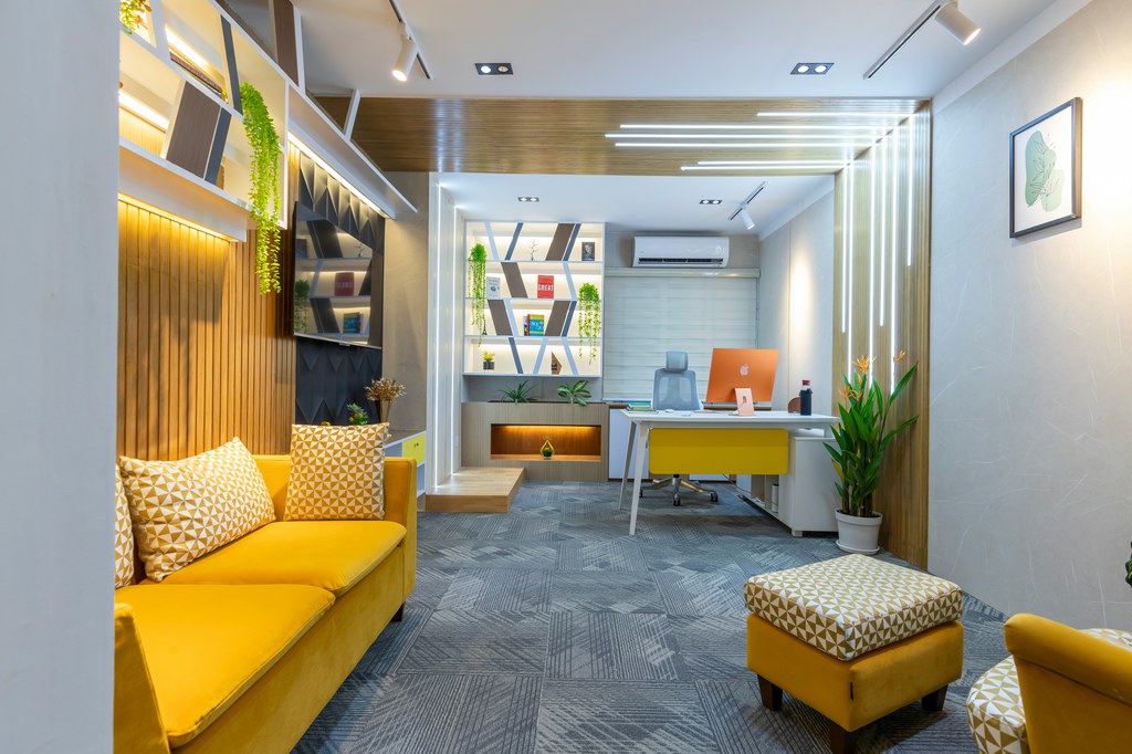 Modern living room office with a yellow sofa, desk, computer, built-in shelving, and plants.
