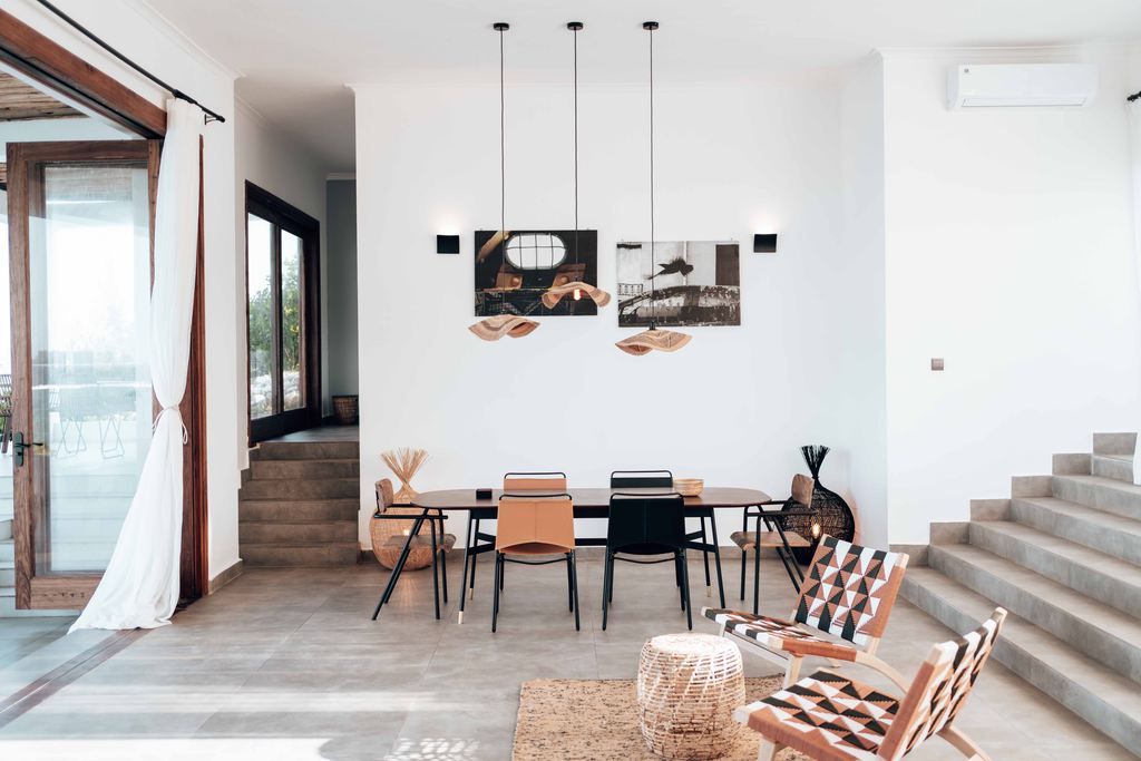 Modern Mediterranean dining area with white walls, warm wood accents, woven pendant lights, minimalist furniture, and a bright open layout.