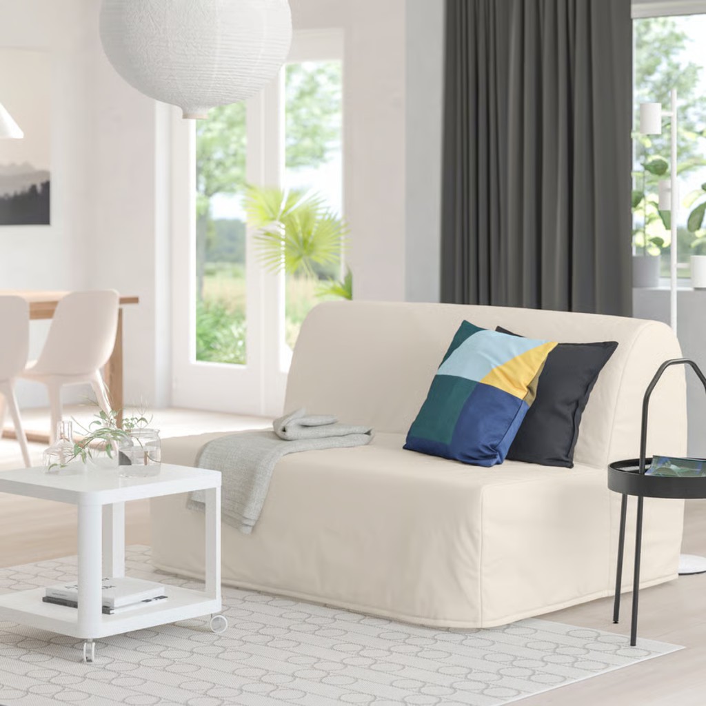 White sleeper sofa with colorful pillows in a bright modern room with large windows, a coffee table, and side table.