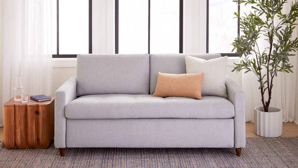 Light gray mid century sleeper sofa with tapered wood legs in a bright living room.