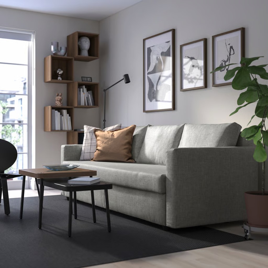Gray sleeper sofa with supportive cushions in a modern living room with coffee tables, wall art, shelving, and a large plant.