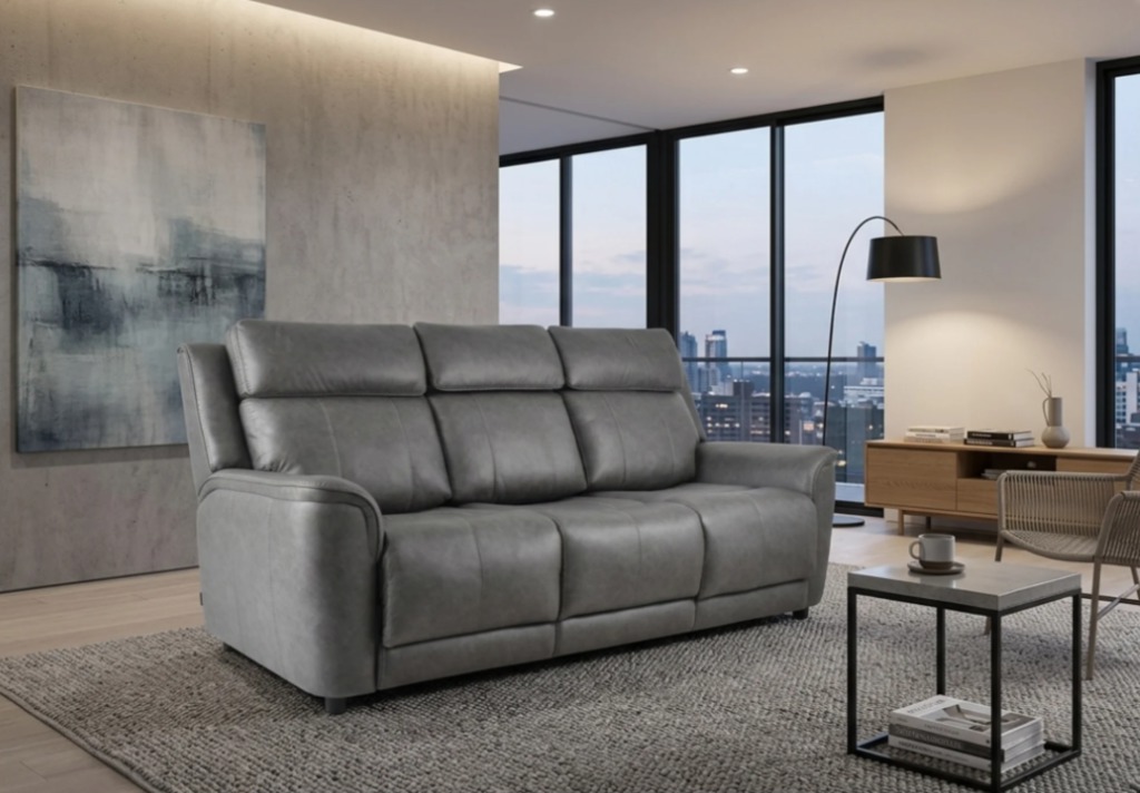 Gray three-seater leather sofa with padded backrests and wide armrests in a modern living room with floor-to-ceiling windows and city views.