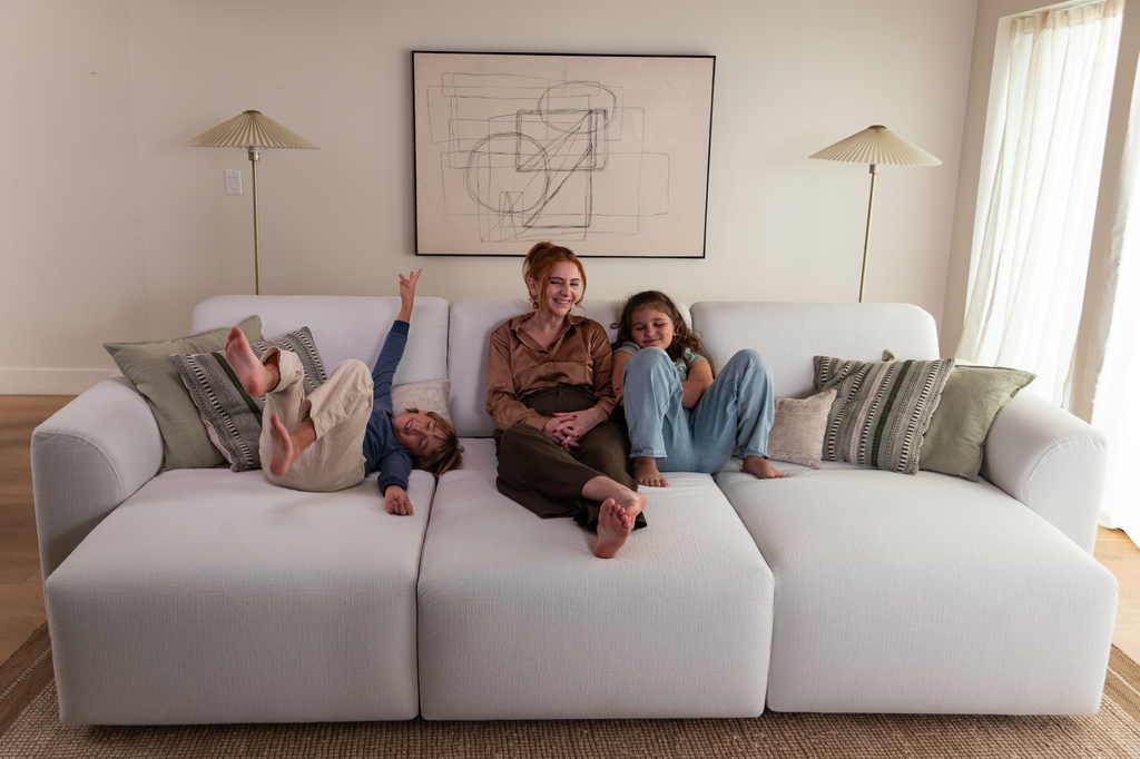Family relaxing together on a large modular white sofa in a bright living room with decorative pillows and a minimalist wall artwork.
