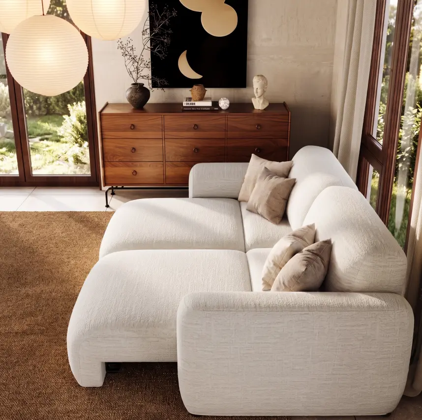 White modular sofa bed with extended seats and beige throw pillows in a sunlit living room with wooden furniture and large windows.