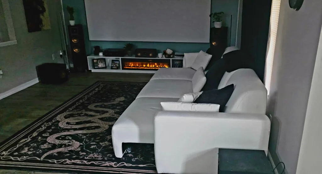White sofa bed facing a projector screen in a small home theater with a media console and floor speakers.